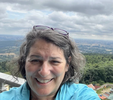 A smiling woman with wavy gray hair and glasses resting on her head takes a selfie outdoors with a scenic landscape of rolling hills, trees, and a cloudy sky in the background. She is wearing a light blue jacket and appears to be on an elevated viewpoint.