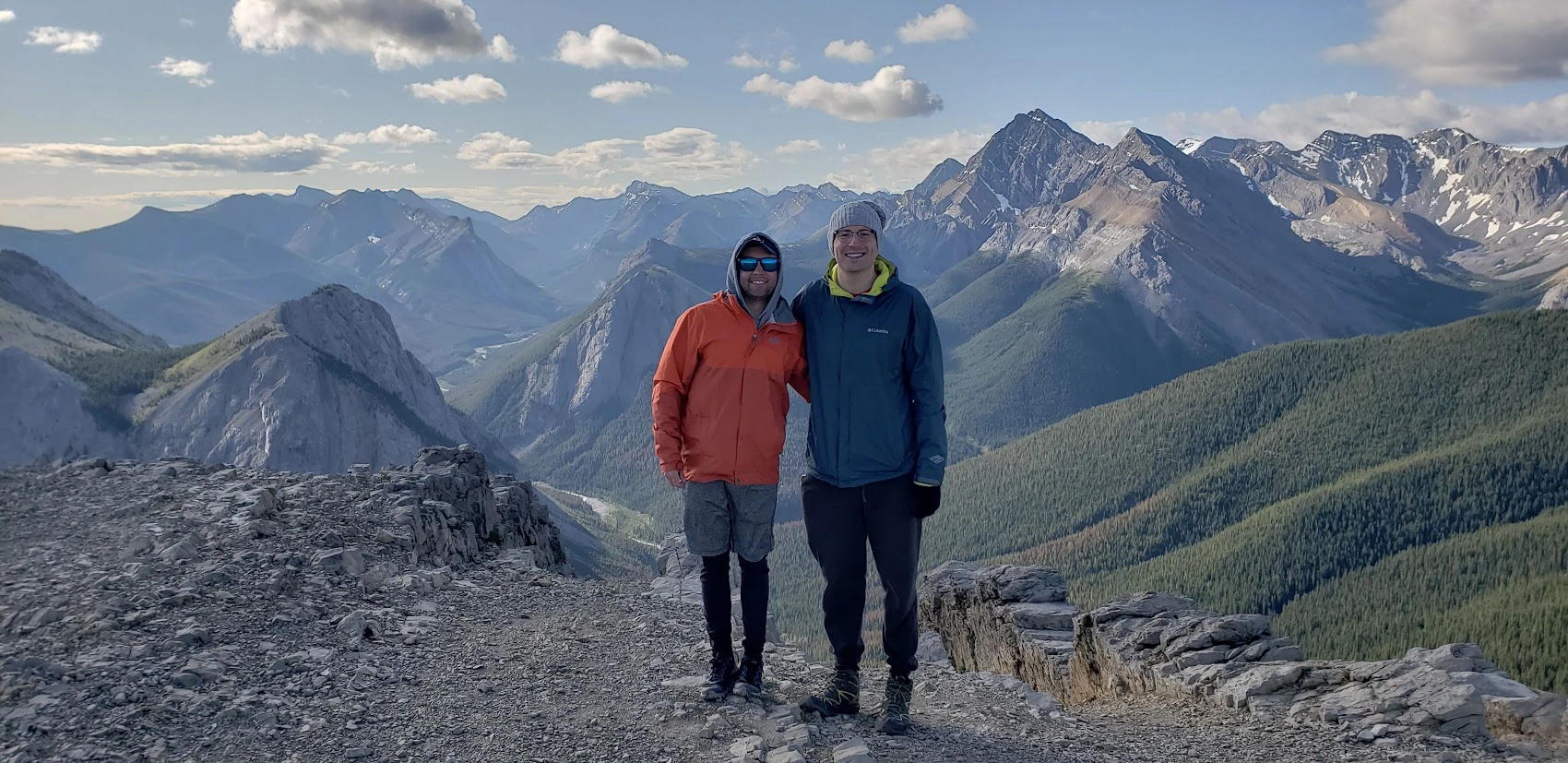 Two men smiling and standing on a rocky mountain ridge with scenic views of the Canadian Rockies in the background.