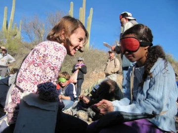 two girls sitting facing each other, one is blindfolded, other people in the background, outside in a desert scene