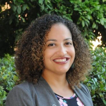 Desiree Vega standing outside under a sunny sky, wearing a gray blazer and floral blouse, greenery in the background