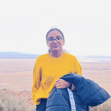 Yamini Sumalatha Bhukya wearing a yellow sweatshirt, holding a jacket, standing outside in a desert lanscape