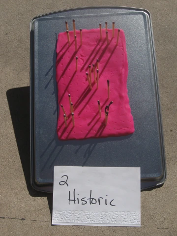 A rectangular piece of pink modeling clay on a baking sheet with multiple wooden matches inserted upright. Some matches have been burned, leaving blackened remnants and small holes in the clay. Shadows from the remaining matches stretch across the surface. A handwritten sign in front reads '2 Historic' in black ink on white paper. The setup is outdoors on a concrete surface.