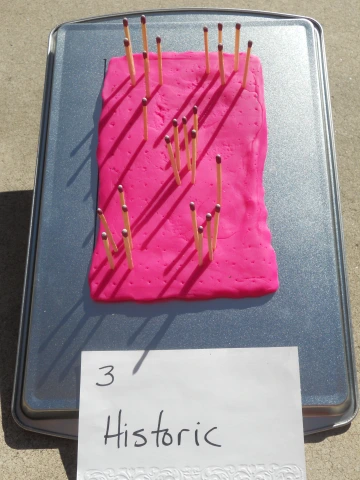 A rectangular piece of pink modeling clay on a baking sheet with multiple wooden matches inserted upright. Shadows from the matches stretch across the surface. A handwritten sign in front reads '3 Historic' in black ink on white paper. The setup is outdoors on a concrete surface.