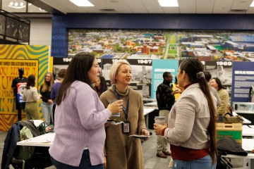 A group of people gathered in an indoor event space, engaging in conversation. Three women in the foreground are holding drinks and plates of food while talking. The background features historical timeline displays, a large aerial photo of a university campus, and an exhibit with bold yellow and blue signage. Other attendees are mingling and interacting.