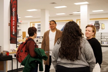  group of four people engaged in conversation inside a retail store. A man in a tan coat and white shirt stands smiling, while three women, one with a red backpack, another with long curly hair, and a third with short blonde hair, listen and interact. The store features technology displays, a 'Back to School' sign, and a refreshment table with drinks in the background.