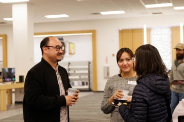 A group of three people engaged in conversation inside a technology retail store. A man wearing glasses, a black jacket, and a patterned shirt holds a cup of coffee while smiling. A woman in a gray plaid dress with a name tag holds a coffee cup and a plate of snacks. Another woman, wearing a navy blue quilted jacket and glasses, is facing them. In the background, a man in a baseball cap stands near the customer service area.