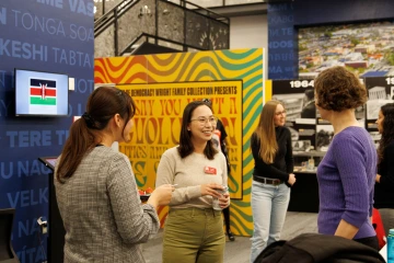 A group of people engaged in conversation at an indoor event. A woman wearing glasses, a beige sweater, and a red name tag holds a cup while smiling. Another woman in a plaid dress holds a plate of strawberries, and a third woman in a purple sweater listens. In the background, attendees are mingling near an exhibit with bold yellow and red signage, a timeline display, and a screen showing the Kenyan flag.