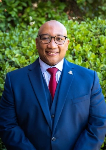 Portrait of Robert Berry, a bald man wearing glasses, a navy blue suit, white dress shirt, and a red tie. He is smiling and standing outdoors in front of green foliage, with a University of Arizona lapel pin on his suit jacket.