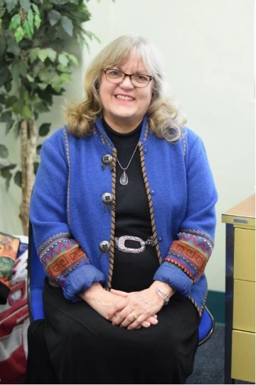 Smiling woman with blonde hair and glasses, wearing a vibrant blue jacket with colorful embroidered sleeves and black dress, seated indoors beside a filing cabinet and a potted plant.