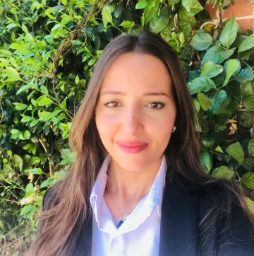 Yousra smiles in a professional headshot taken outdoors in front of green ivy. She wears a white collared shirt, a black blazer, and a delicate necklace, with long brown hair and natural makeup.