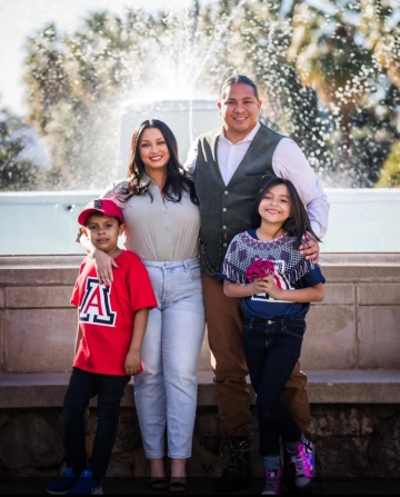 A smiling family of four stands in front of a large water fountain. The mother and father are standing close together in the middle, with the father’s arm around their daughter on the right and the mother’s arm around their son on the left. The young boy is wearing a red University of Arizona shirt and hat, and the girl is holding flowers and wearing sparkly boots. Palm trees and sunlight are visible in the background.