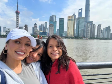 A woman and two smiling children pose for a selfie by the river in front of the Shanghai skyline. The woman wears a white baseball cap and gray top, and the girls are dressed in casual hoodies. One girl playfully makes bunny ears behind the other. Modern skyscrapers, including the Oriental Pearl Tower and Shanghai Tower, rise behind them under a partly cloudy sky.
