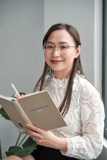 Chi Nguyen sits beside a window, smiling warmly while holding an open beige planner titled ‘THE PRESENT DAY planner.’ She wears a white lace blouse with intricate circular patterns and clear-framed glasses. A pen is in her hand, and a green leafy plant is partially visible in the background.