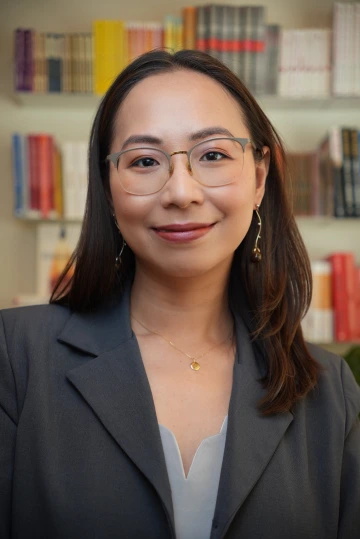 Professional portrait of Chi Nguyen smiling in front of a blurred background of colorful bookshelves. She wears glasses, dangling earrings, a delicate necklace with a round pendant, and a dark blazer over a light-colored top. Her hair is neatly styled, and she appears confident and approachable.