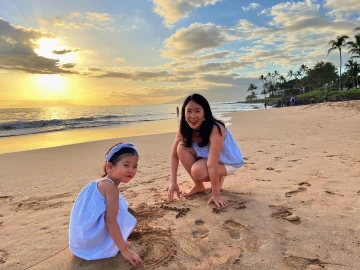 “Dr. Sung Eun Jung and her young daughter play in the sand on a beach during sunset. Both are smiling and wearing light blue outfits. The sun is setting over the ocean, casting a golden glow across the sky, while palm trees and a few people are visible in the distance along the shoreline.