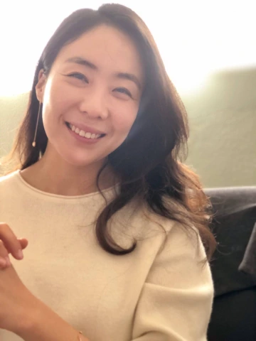 A warm and softly lit portrait of Dr. Sung Eun Jung smiling at the camera. She has long, dark hair and is wearing a cream-colored sweater with a delicate drop earring.