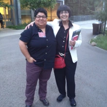 Cindy Cruz stands outdoors beside philosopher Maria Lugones. Cindy wears a black button-up shirt and maroon pants; Maria wears a white blazer over a black outfit and carries a red purse and notebooks. They smile with arms around each other.