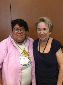 Cindy Cruz, wearing glasses, a light pink shirt, and a printed t-shirt, stands next to Sonia Saldivar-Hull, who is dressed in a navy dress with a beaded necklace. Both are smiling in front of a wooden wall, wearing conference name badges.