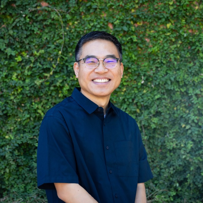 ahn headshot, wearing a blue shirt, eyeglasses, greenery in the background