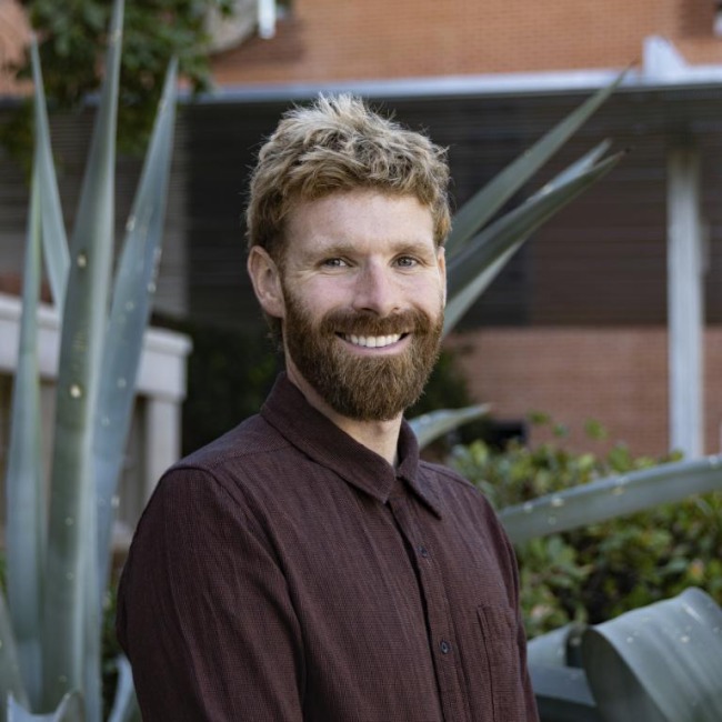 jesse stipek headshot, dark blonde hair, facial hair, wearing a brown button up shirt, standing outside with greenery in the background