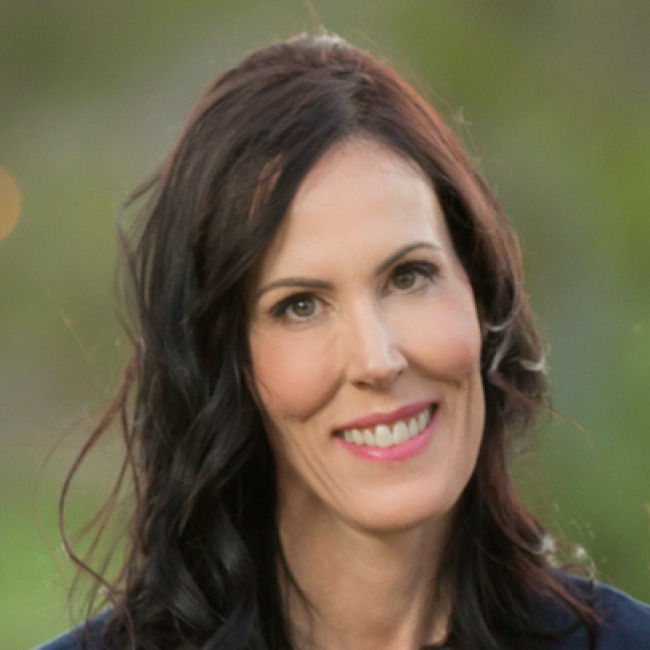 Portrait of a woman with long, wavy dark brown hair, wearing a navy blue top. She is smiling softly and standing outdoors with a blurred green background.