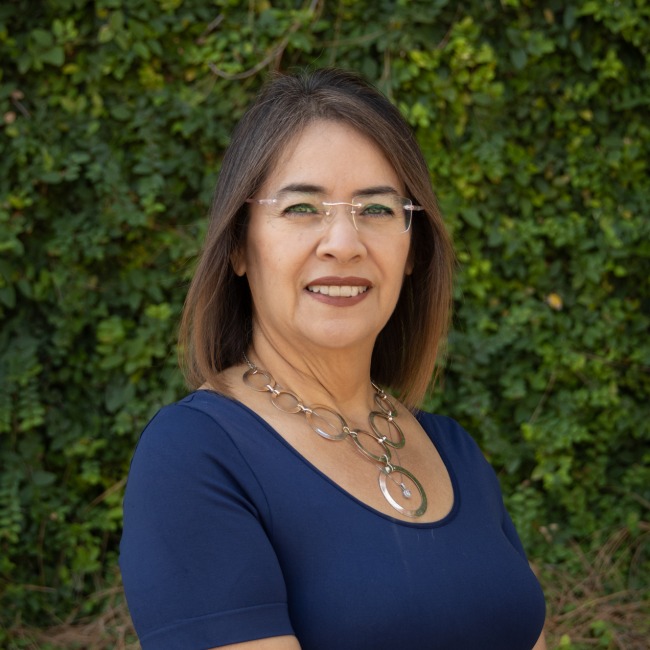 a woman with shoulder-length brown hair, wearing glasses, a navy blue top, and a statement necklace with large circular links. She is smiling and standing outdoors in front of a lush green ivy-covered wall