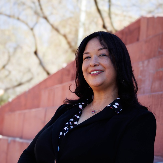 Professional portrait of Dr. Regina Deil-Amen, smiling while wearing a black blazer over a patterned blouse and a silver necklace with a red gemstone. She is standing outdoors in front of red stone steps, with blurred tree branches in the background.