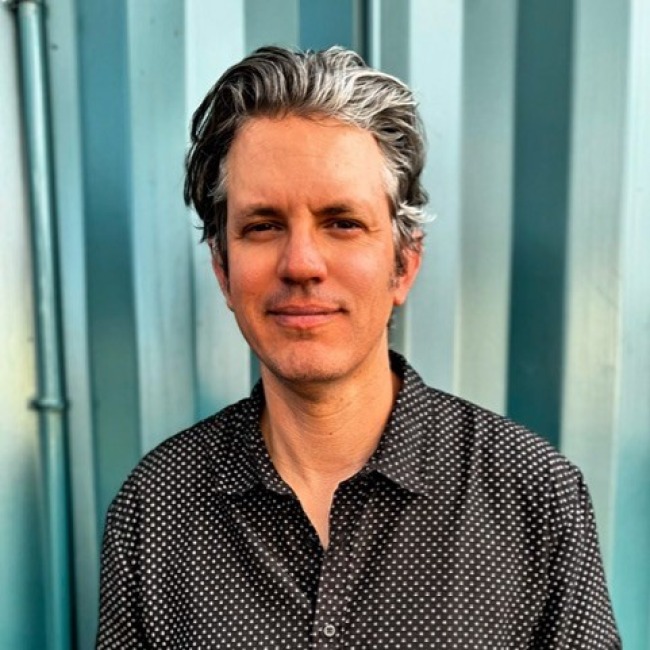 Portrait of Alex Lynch standing in front of a teal metal wall, wearing a black button-up shirt with white polka dots, and smiling slightly at the camera.
