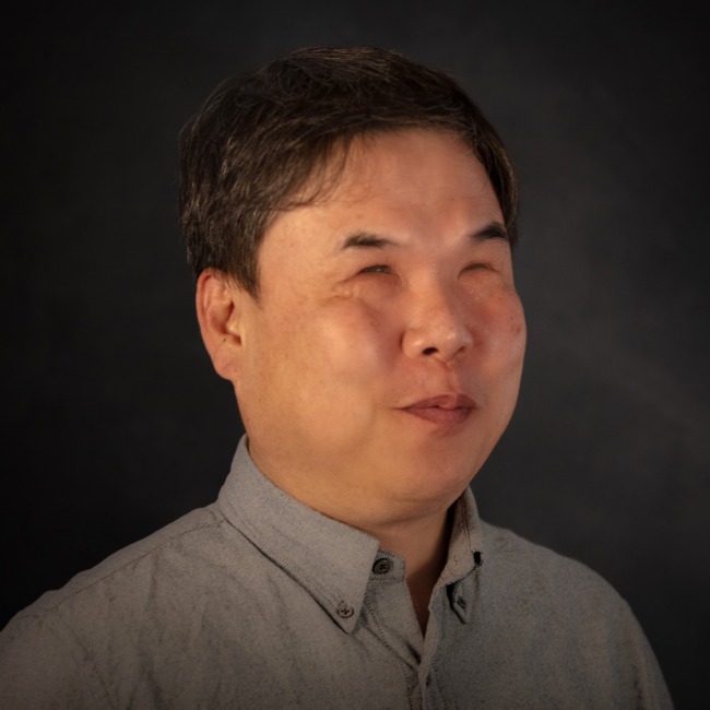 Portrait of a man with short dark hair wearing a button-up shirt against a dark background, smiling slightly.