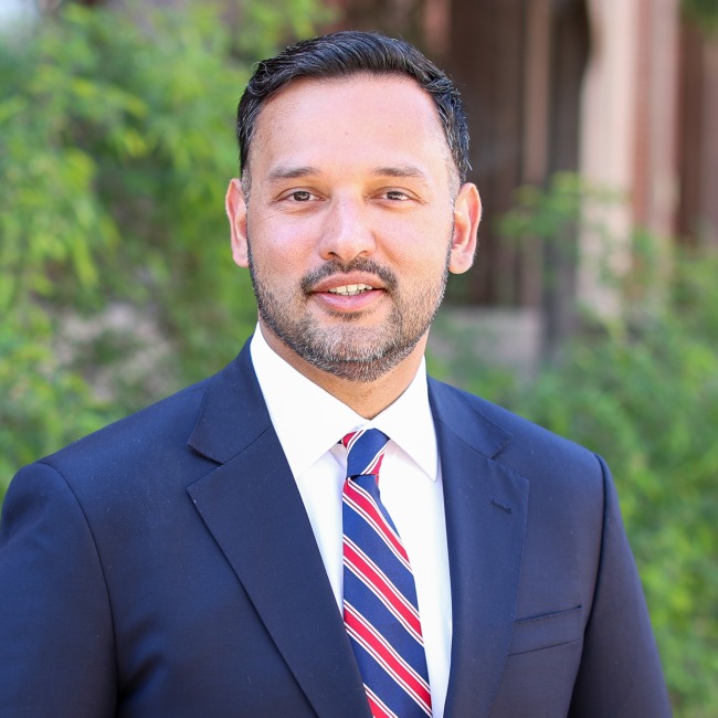 A professional outdoor portrait of Daniel Saud wearing a navy blue suit, white dress shirt, and a red, white, and blue striped tie.