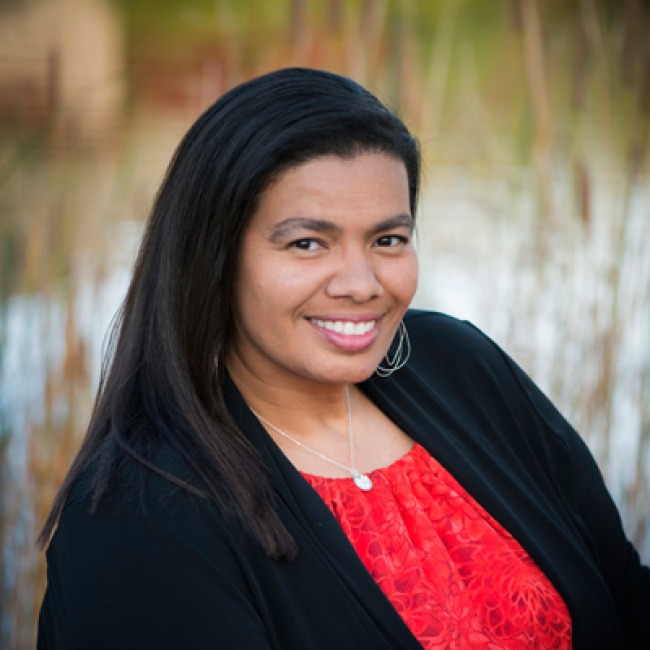 Professional outdoor portrait of a smiling woman with long dark hair, wearing a black cardigan over a bright red textured blouse. She is posed slightly angled to the side in front of a natural background with soft-focus tall grass and water.