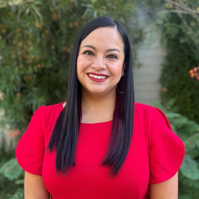 Karla Silva stands outdoors in front of green foliage, smiling warmly at the camera. She is wearing a bold red dress with short sleeves and has long, straight black hair.