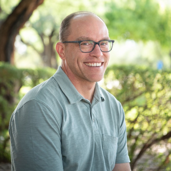Outdoor portrait of a smiling man with glasses and short-cropped hair wearing a light green polo shirt. He is seated in front of sunlit trees and greenery, with soft, natural light creating a bright and welcoming atmosphere.