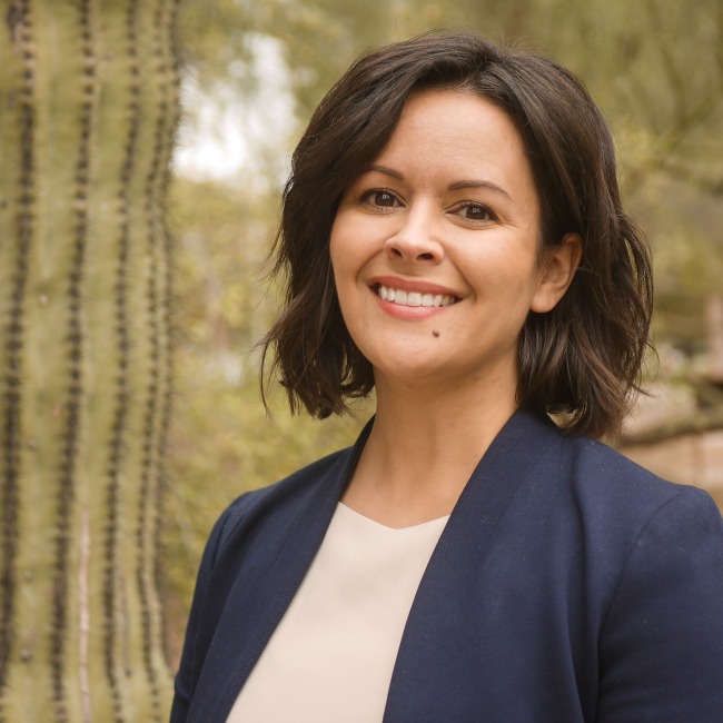 Professional headshot of a smiling woman with short dark hair wearing a navy blazer over a cream blouse. She is standing outdoors next to a large saguaro cactus, with a blurred desert landscape in the background.