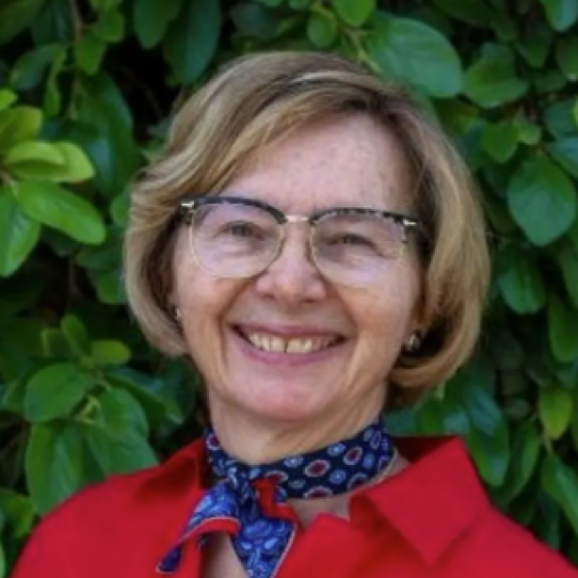 Sharon Wisniewski stands smiling in front of a backdrop of lush green leaves. She is wearing glasses, a bright red blouse, and a patterned blue scarf tied around her neck.