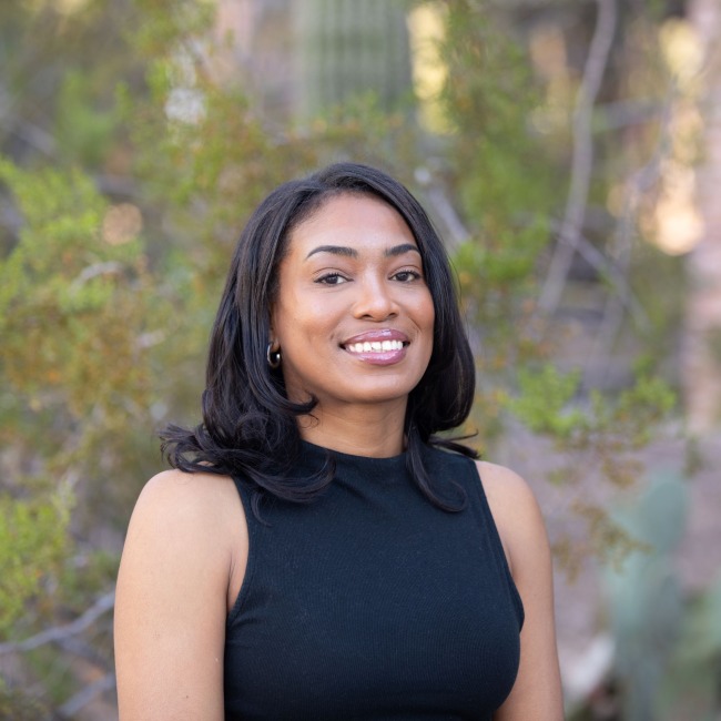 Crystal Govan smiling outdoors in front of desert vegetation, wearing a sleeveless black top with loose, shoulder-length hair.