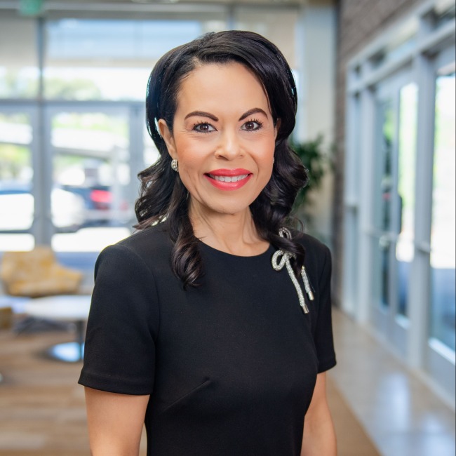 Portrait of Marla Franco smiling indoors, wearing a black dress with a rhinestone pin, standing in a brightly lit hallway with glass doors and modern furniture in the background.