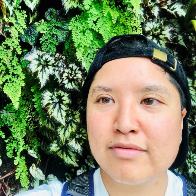 Person wearing a backward black cap and white shirt standing in front of a wall covered with vibrant green and variegated foliage.