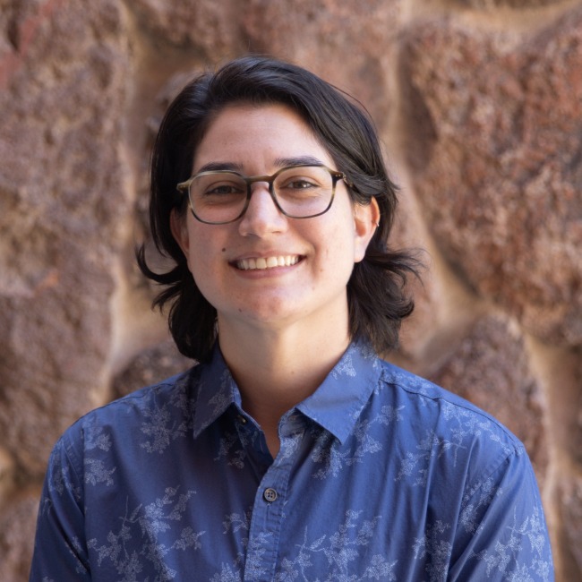 Portrait of Michele Stathatos smiling in front of a stone wall, wearing glasses and a blue patterned button-down shirt